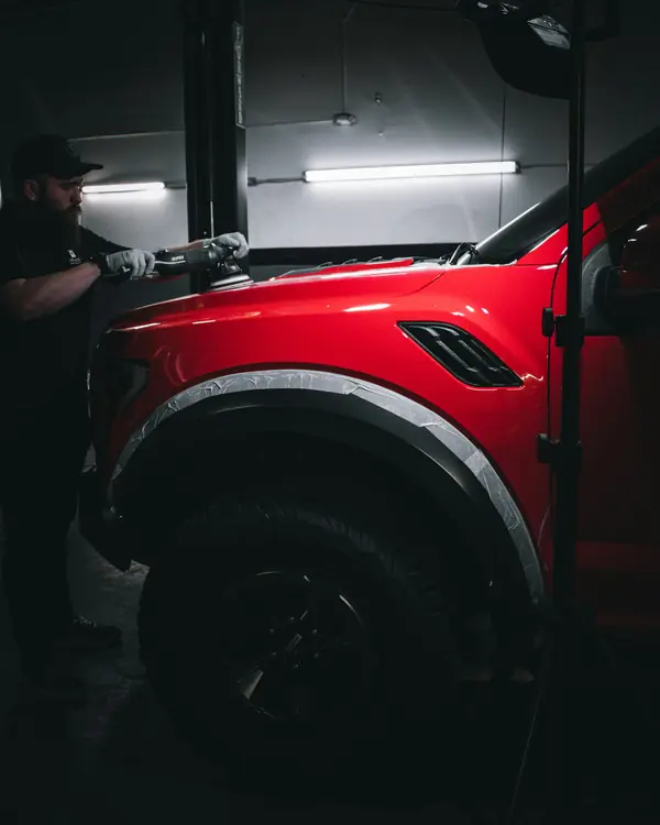 A detailer machine-polishing the bonnet of a red truck beside an inspection lamp
