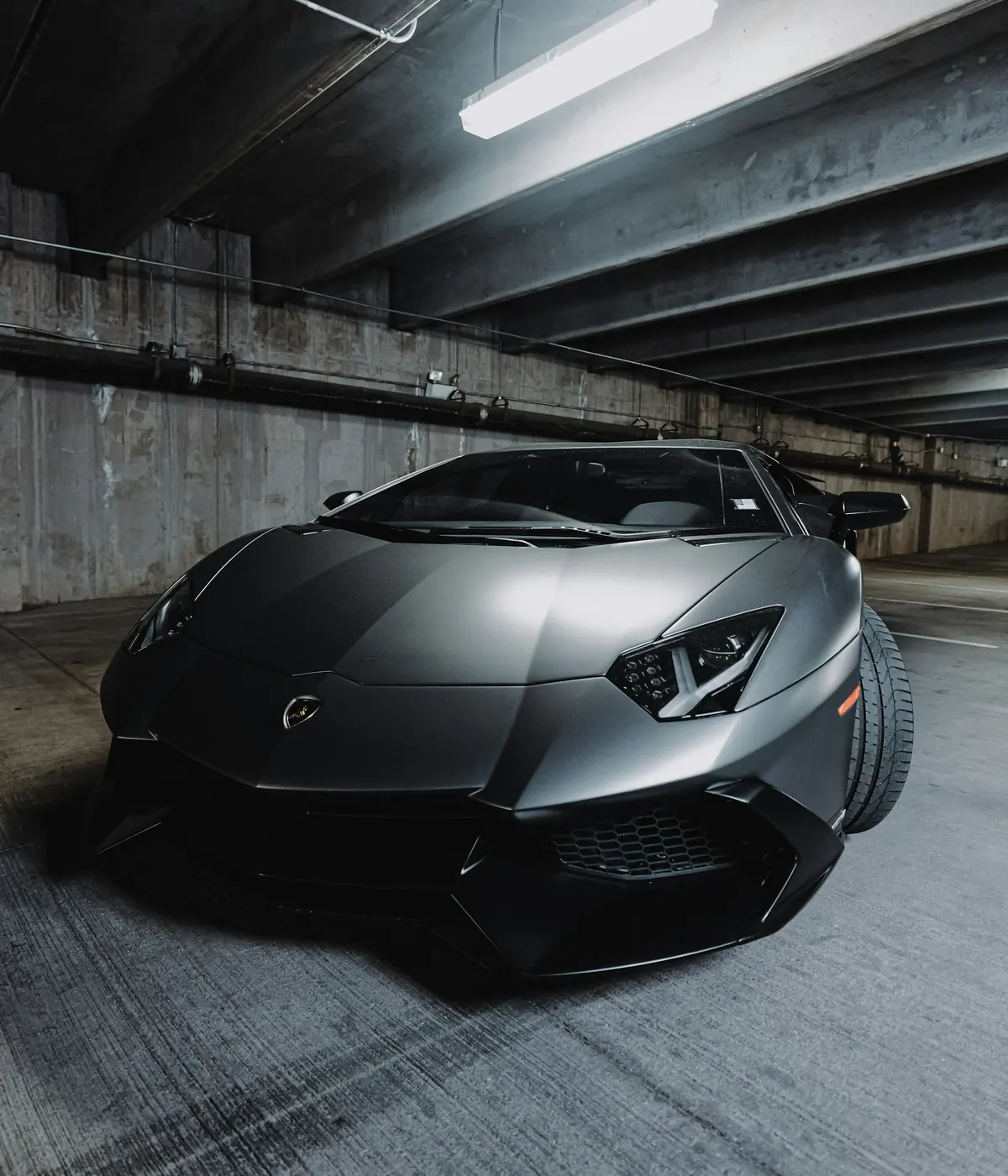 A matte grey supercar parked under a strip light in a concrete garage