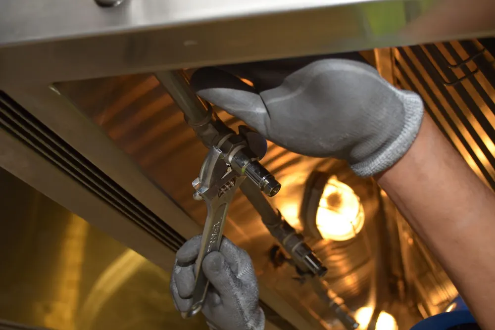 Gloved hands turning a wrench on a pipe fitting under a hood